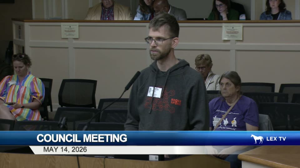 A man in a charcoal Broke Spoke Community Bike Shop hoodie speaks at the LFUCG public-comment podium with a visitor badge clipped to his chest, May 14, 2026.