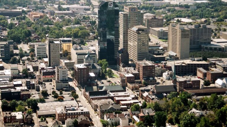 An aerial daytime view of the downtown Lexington skyline, featuring a cluster of tall office buildings surrounded by smaller