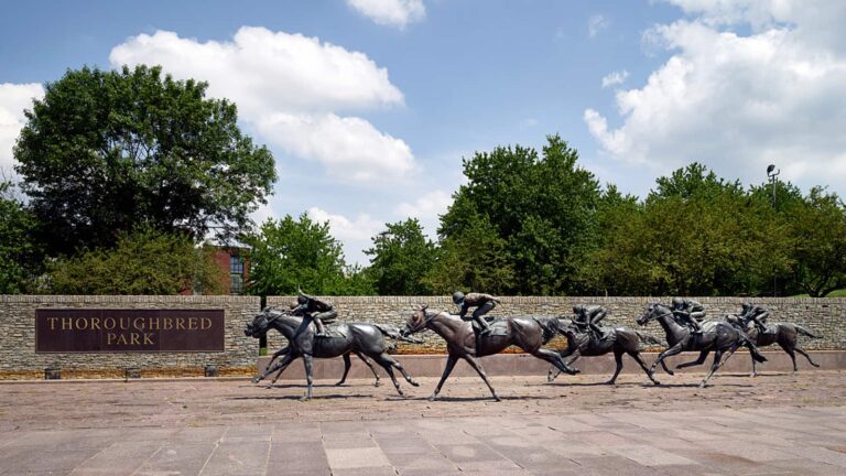 Life-size bronze sculptures of jockeys racing thoroughbred horses at Thoroughbred Park, set against a low stone wall, lush
